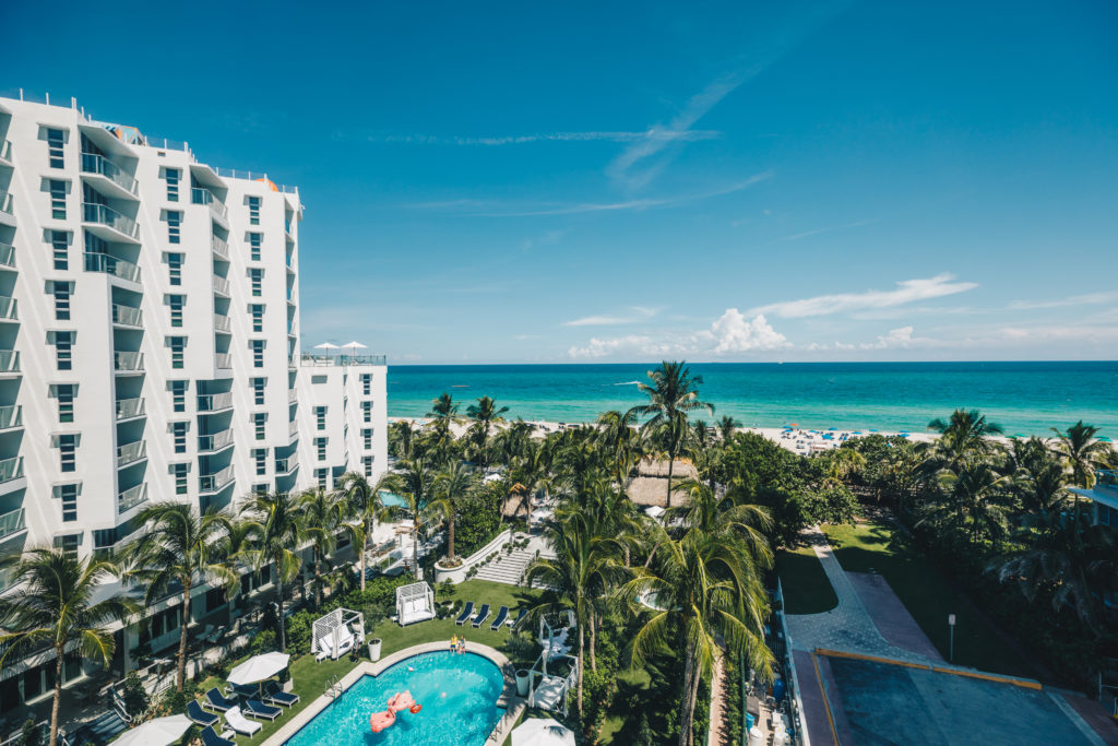 Aerial view of the Cadillac Hotel & Beach Club property in Miami Beach, from the pool surrounded by palm trees to the ocean in the background