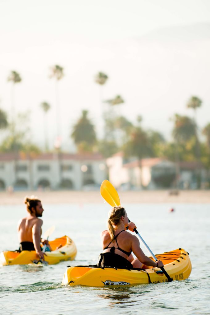 Two people in yellow kayaks in Santa Barbara. Palm trees and Spanish Colonial style buildings in the background.