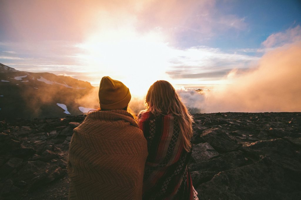Man and woman wrapped in blankets on top of a misty mountain watching the sunrise.