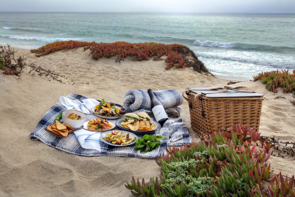 Plates of food and a bottle of wine set up on a fleece blanket on the beach, next to a picnic basket. Picnic at The Sanctuary Beach Resort.