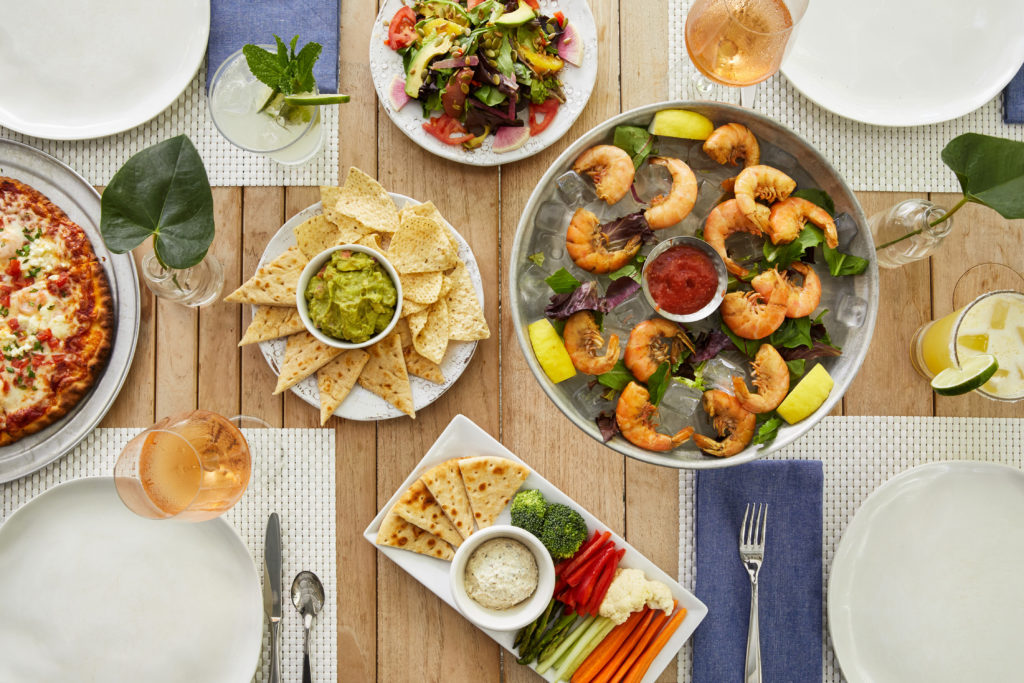 A variety of food on a wooden table at The Grove Kitchen & Bar at Parrot Key Hotel & Villas. Flat lay of shrimp cocktail, hummus plate, chips and guacamole, cocktails, and place settings.
