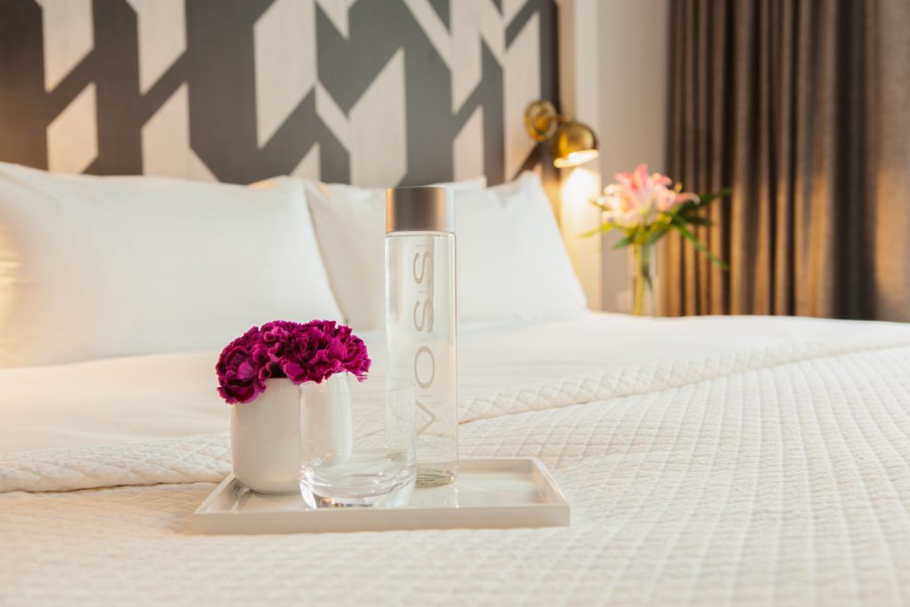 A tray of flowers, a glass, and bottle of Voss water on a guest room bed at The Tillary Hotel in Brooklyn